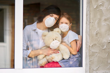 Mother with daughter child in mask looking from window, coronavirus.