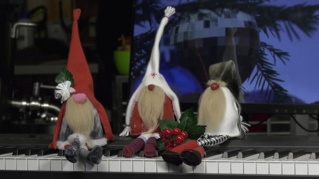 A group of cheerful dwarves sits on the piano against the background of a monitor with a New Year's toy. Homemade figurines made of fabric are placed on the piano keys. Camera movement panorama.