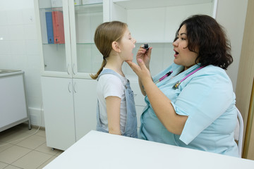 The girl came to the pediatrician with an otoscope. Smiling otolaryngologist doctor looking at a child while examining a child's throat with a tongue depressor.
