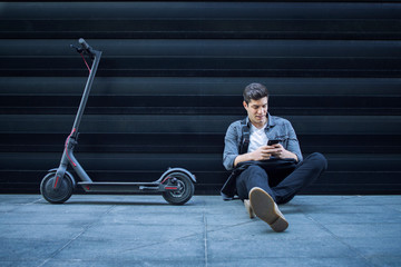 Shot of young caucasian hipster using smart phone while siting on the ground next to his electric...