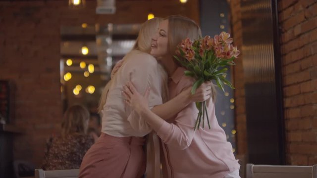 Handheld shot of happy woman meeting her mother in cafe and gifting her bouquet of flowers on mothers day or her birthday. She is smiling and kissing her on cheek, then sitting down at table to chat