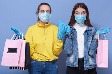 Closeup portrait of two young girls wearing casual outfits and protective masks and gloves, holding shopping bags, spreading hands aside, con not resist shopping during . Coronavirus, Civid19 concept.