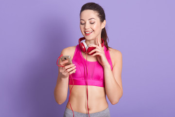Indoor portrait of cheerful beautiful young female smiling sincerely, having pleasant facial expression, looking at device screen, holding smartphone, wearing sport clothes. Fitness trainer concept.