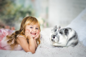 Portrait of cute smiling blonde 3 years old girl in pink dress laying on bed with funny furry gray and white rabbit. Rabbit washing his paw and happy child. Easter rabbit and small kid. Indoor
