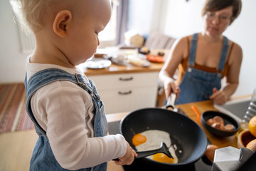A child in the interior of a stylish kitchen with his mother prepares breakfast, fries eggs in a pan on the stove. Helping and learning.