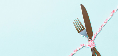 Diet and healthy eating concept with crossed fork and knife and curled measuring tape on blue background. Top view of weightloss with copy space