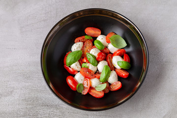 Caprese salad with mozzarella, tomatoes and basil leaves in black bowl on grey background. Healthy vegetarian lunch or dinner. Italian traditional food. Top view. 