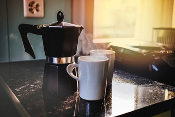 Preparing fresh coffee in moka pot on electric stove in the early morning. Pouring hot steaming coffee into two white mugs.