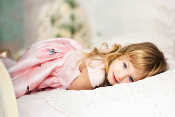 Obraz na płótnie Canvas Portrait of beautiful cute little 3 years old blond girl with blue eyes lying on bed in pink dress. Moment of relaxation for active child. Kid is bored indoors