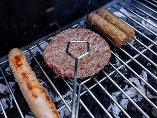 Sausages and beef burger grilling over hot coals with selective focus on an unidentifiable chef flipping the smoking burger with a set of metal tongs