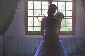 Beautiful bride in white wedding dress standing in her bedroom and looking in window.
gorgeous happy luxury stylish brunette bride near a window on the background of vintage room.