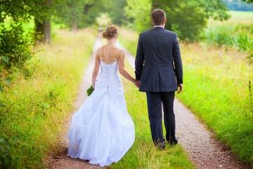 Back view of bride in white dress and groom in suit holding each others hands outdoors
Happy bride and groom walking in nature on wedding day. Wedding couple in love, newlyweds, copy space, no face