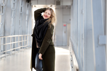 Stylish fashion portrait of blonde woman. Posing in the city. Beautiful girl in autumn green coat and black scarf poses in an overhead pedestrian crossing.