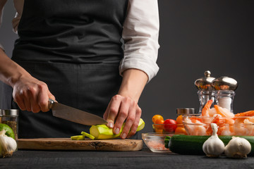 A professional chef cuts zucchini to make a salad with seafood, shrimp. On a gray background with ingredients.