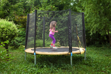 Garden trampoline with little child moving inside. Trampolin in the garth with little girl jumping. Fun on garden trampoline in summer day. Recreational trampoline with a safety net enclosure.