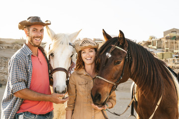 Happy couple having fun with horses inside stable - Young farmers sharing time with animals in corral ranch - Human and animals relationship lifestyle concept
