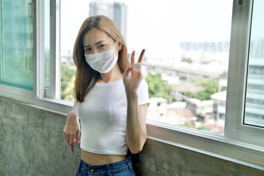 Social distancing concept. Young Asian lady with face mask showing victory gesture during staying and working at home in quarantine period. Self quarantine. Covid 19 outbreak pandemic.