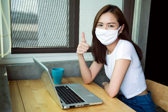 Social distancing concept. Young Asian lady with face mask and hand thumb up working at home during quarantine period. Self quarantine. Corona virus Covid 19 outbreak pandemic.