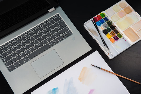 Graphic designer's desk table, laptop, watercolors palette and sheet for painting on a dark wooden table.
