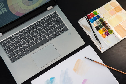 Desk table, laptop, watercolors palette and sheet for painting on a dark wooden table, workplace of a designer.