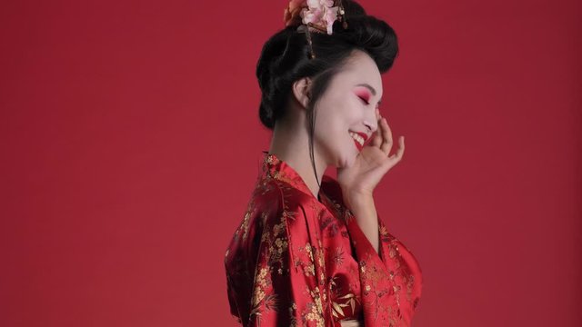 A pretty nice young geisha woman in traditional japanese kimono is posing while turning to the camera isolated over red background