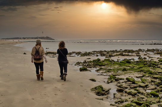 Back view of two women walking on a beach of Cadiz called Zahora.  Selective focus on the rocks covered with moss. Trafalgar lighthouse and beautiful sunset in the background.