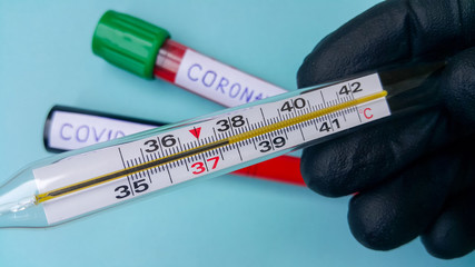 Obraz premium A thermometer showing a high temperature in the doctor's hand. In the background, without focus, there are two vacuum tubes with blood. Text of coronavirus and covid-19. The concept of infection.