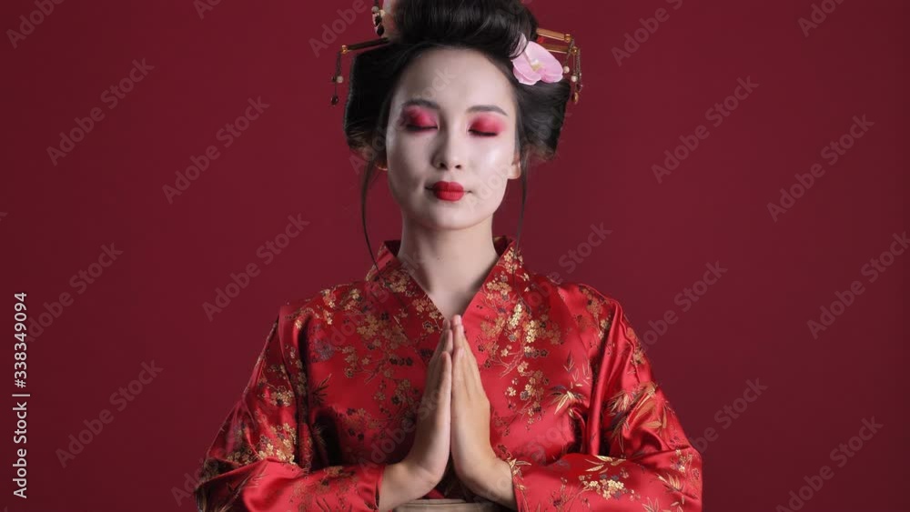 Sticker A calm young geisha woman in traditional japanese kimono is holding hands together like a praying pose isolated over red background