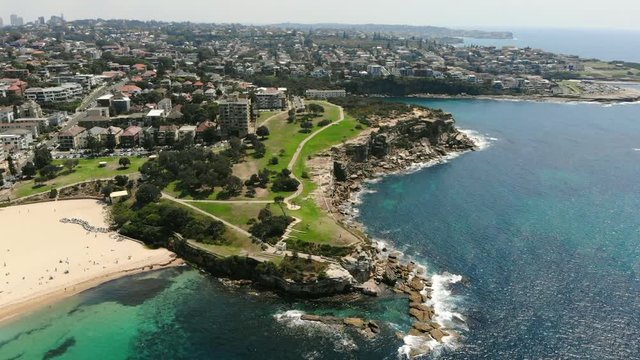 Sydneys Coogee beach, drone slowly moving forward start of Coogee to Bondi walk. Grass banks with clear water and beach surrounding it with city in the background