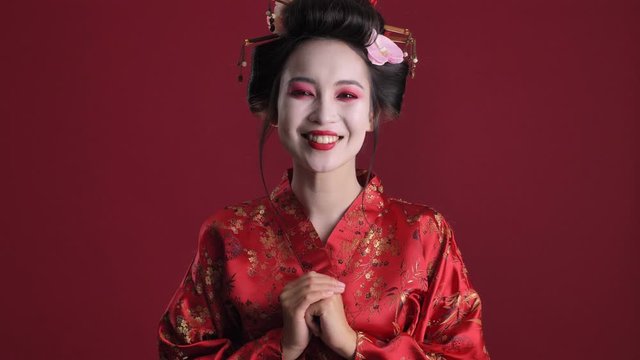 A happy joyful young geisha woman in traditional japanese kimono is winking while posing isolated over red background