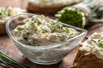 A bowl of homemade cream cheese spread with chopped chives surrounded by bread slices with spread and a bunch of freshly cut chives - obrazy, fototapety, plakaty
