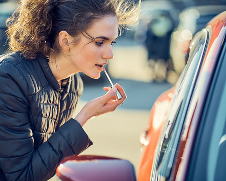 Young attractive woman looks in the side window of a car and using it as a mirror, paints her lips. makeup in the car and on the street.