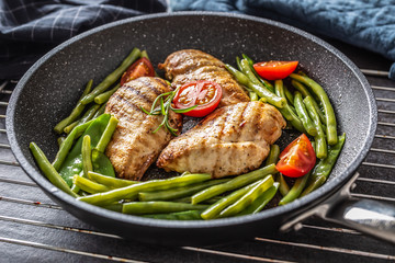 Healthy recipe of grilled chicken, green vegetable and tomatoes on a dark nonstick pan placed on metalic grid and dark background