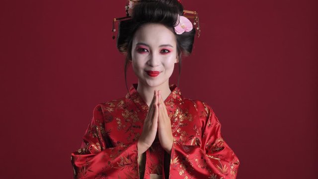 A positive attractive young geisha woman in traditional japanese kimono is doing traditional japanese greeting isolated over red background
