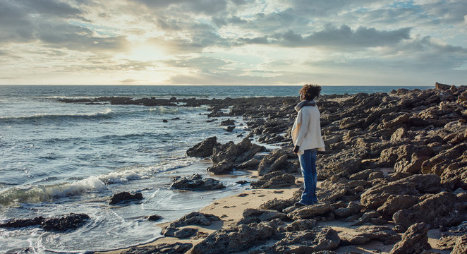 Side view of a black woman standing on a rock and looking at the horizon in Zahora beach, Cadiz, Andalusia, Spain. Beautiful sunset.  