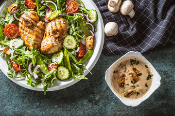 Top view of grilled chicken and mushrooms with salad, cucumber, cherry tomatoes, red onion, with a creamy mushroom sauce in a separate bowl on dark background