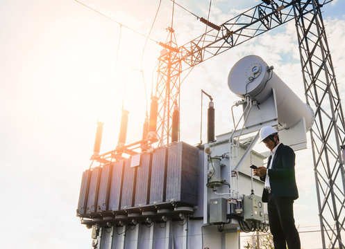 A low angle image of a businessman wearing a black suit, standing looking at a large power transformer with blue sky to be background, Concept about business people who want to invest in energy