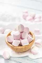 Portrait of a wooden bowl full of pink and white marshmallows with some scattered around on a white...