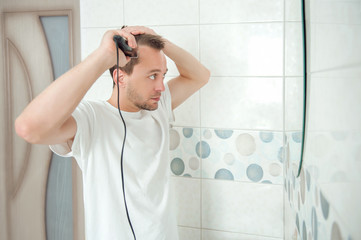 young attractive man makes a haircut to himself in front of a mirror in the bathroom with a hair clipper. man cuts his hair