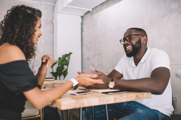 Cheerful male and female friends enjoying live communication during free time in cafeteria interior, youthful African American hipster guys smiling during positive date in public coffee shop