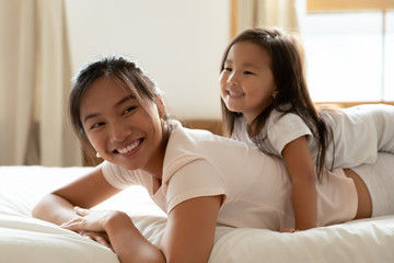 Obraz na płótnie Canvas Vietnamese family play while lying on bed in warm cozy bedroom, little adorable daughter and asian beautiful mother relish time together wear comfortable pyjamas woke up enjoy new day feels overjoyed