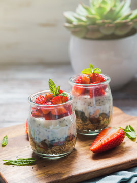Yogurt granola parfait with strawberries and chia seeds. Healthy single portion breakfast served in glass cups on wooden board with green napkin. Natural wooden table background