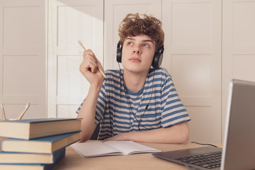 A nice teenage boy in headphones uses the laptop and learns at the desk in his room. He takes notes with a pencil. Distance learning because of the epidemic coronavirus