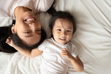 Asian single mother little daughter lying down together on bed white bedding smiling looking at camera feels joyful, above close up view. Happy motherhood, next generation, beautiful family portrait