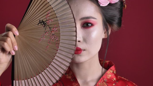 A close-up view of a joyful young geisha woman in traditional japanese kimono is posing with wooden hand fan isolated over red background