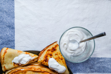 Top view on a stack of rosy freshly baked pancakes on a brown plate. Nearby is a glass transparent cup with white sour cream and a metal spoon. Tasty breakfast from the village.
