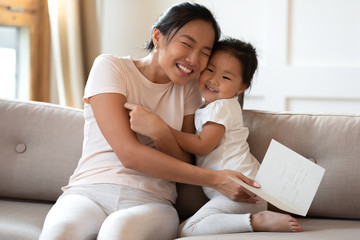 Grateful happy asian mom received birthday present handmade post card read wishes feels appreciative cuddles her small attentive cute daughter. Life event, love, care, Mother Day celebration concept