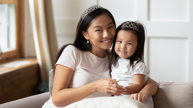 Beautiful mother cuddles adorable preschool daughter, asian family wearing casual clothes and crown on heads photo shooting seated on couch in living room. Happy motherhood, unconditional love concept