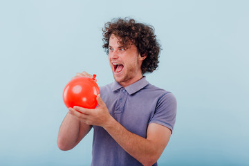 Crazy looking young guy with curly hair holding half inflated red balloon and laughing at voice sounding after inhaling helium gas on blue background