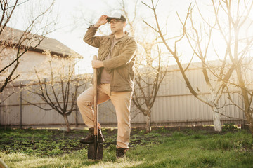 Adult thoughtful man posing outside. Hold one feet on shovel. Beautiful sunny spring day on backyard. Keep one hand on cap.